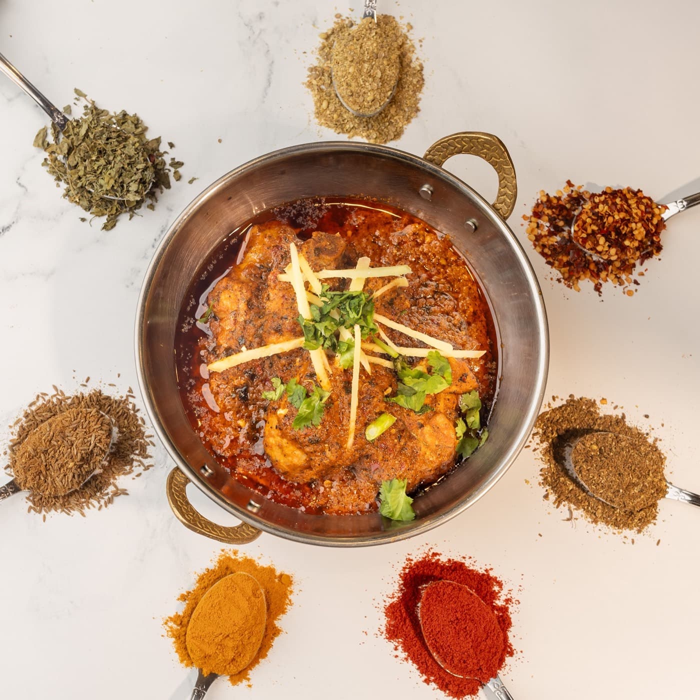 Chicken karahi in a steel karahi, surrounded by spoons of haldi, lal mirch, zeera and dhania